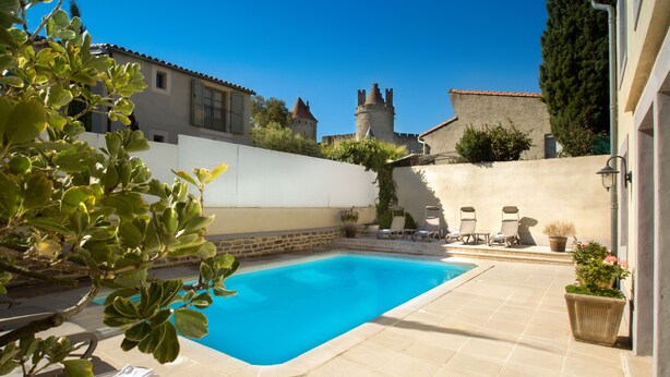 Uma piscina com espreguiçadeiras, uma parede de pedra e um castelo ao fundo.