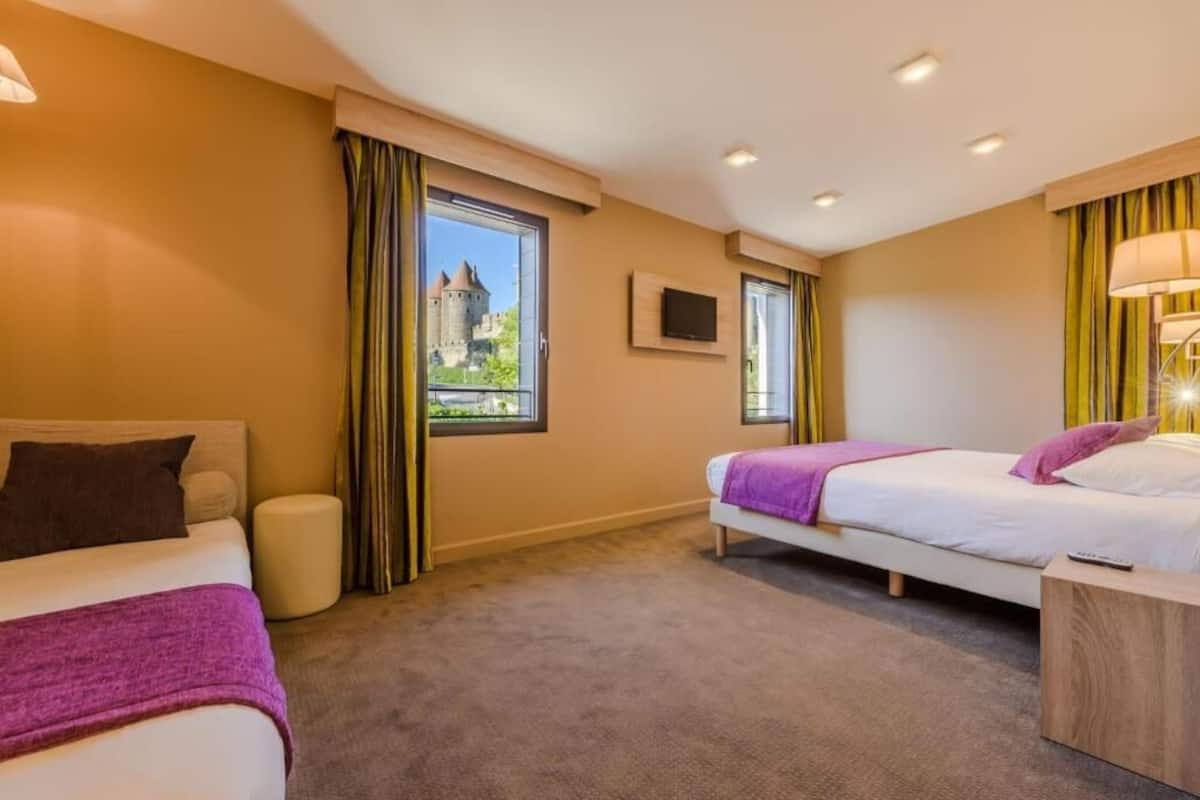 A hotel room with two beds, a TV, a window with a view of a castle, and a lamp.