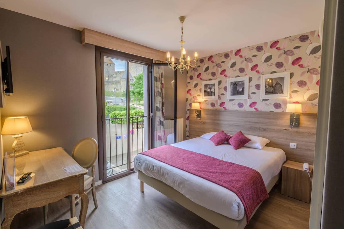 A hotel room with a large bed, a desk with a chair, a balcony with a view of greenery, and a wall with framed pictures.