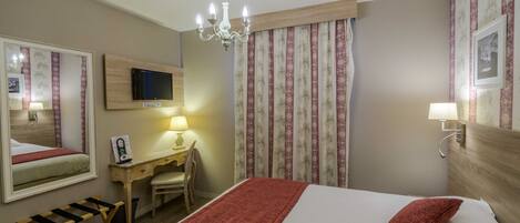 A hotel room with a bed, a desk, a chair, a TV, a chandelier, and a window with curtains.