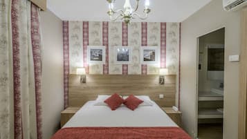 A hotel room with a large bed, a wooden headboard, a chandelier, and a bathroom visible through an open door.