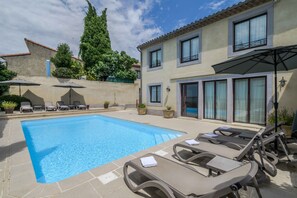 Une piscine entourée de chaises longues et de parasols, bordée d’un bâtiment doté de grandes fenêtres et d’un toit recouvert de tuiles.