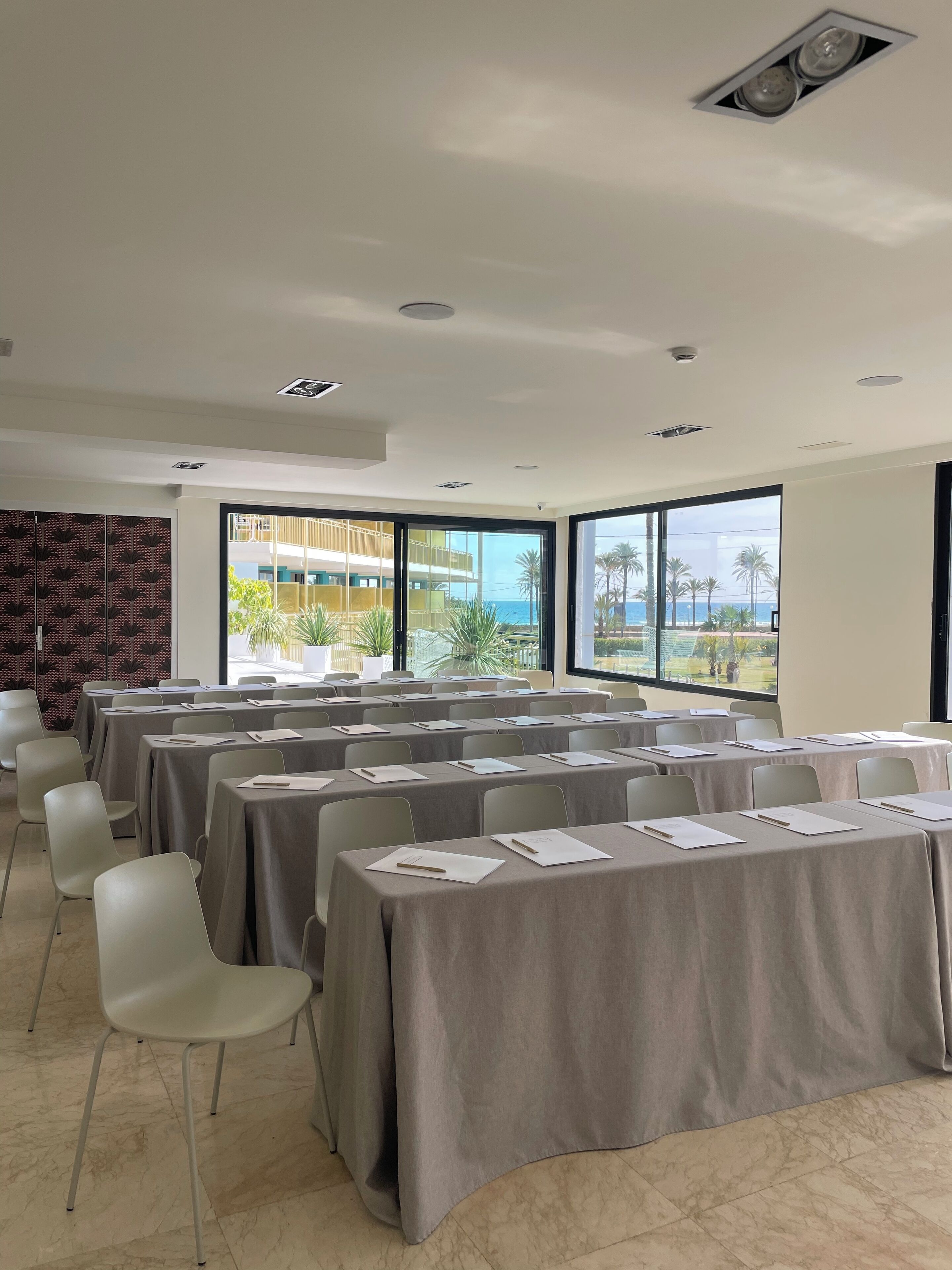 Un salón de conferencias con mesas dispuestas para reuniones, amplias ventanas que ofrecen vistas a palmeras y un edificio, y un techo moderno con iluminación empotrada.