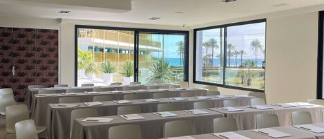 Un salón de conferencias con mesas dispuestas para reuniones, amplias ventanas que ofrecen vistas a palmeras y un edificio, y un techo moderno con iluminación empotrada.