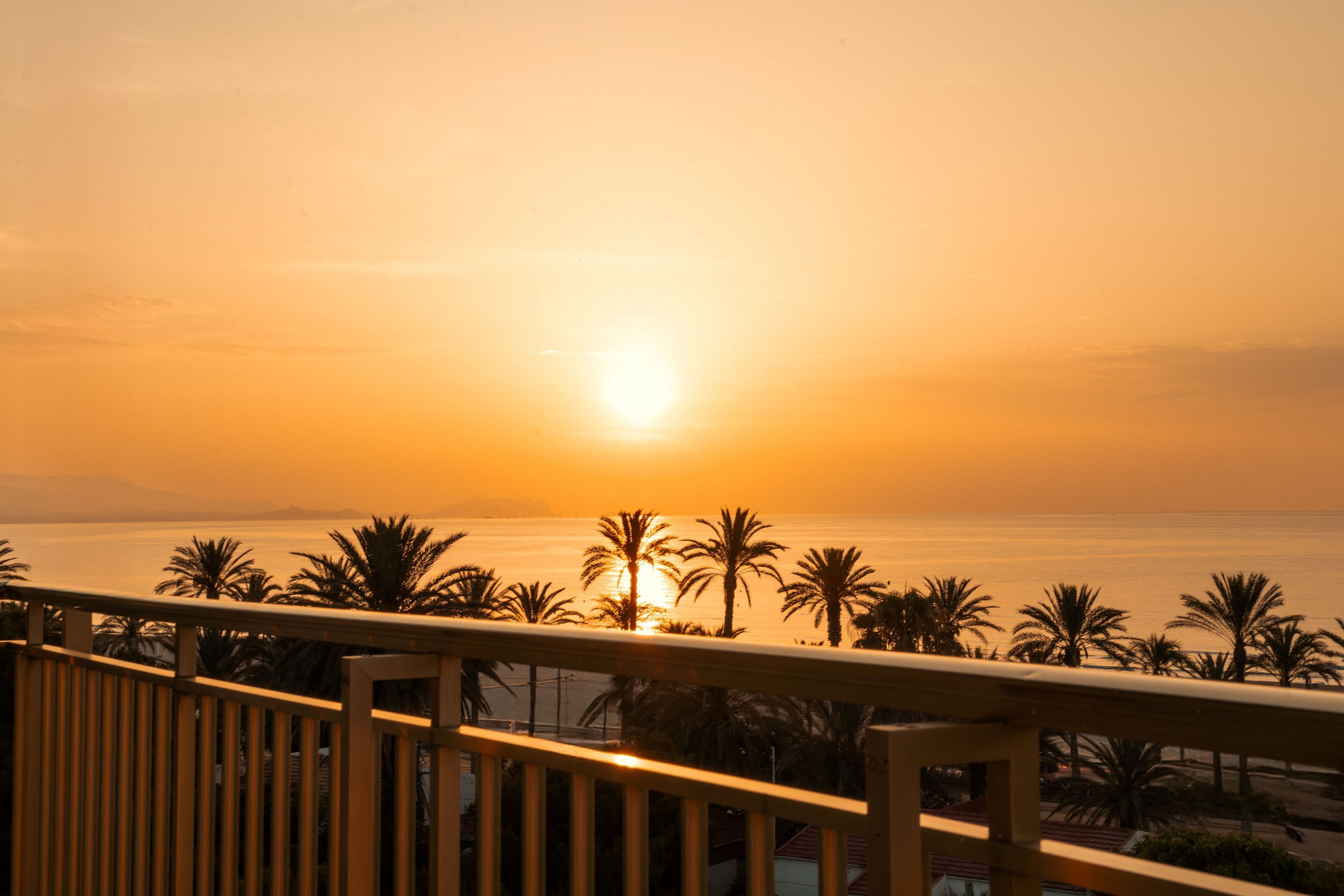 Un balcón con vista a la puesta de sol sobre el mar, palmeras y montañas distantes.