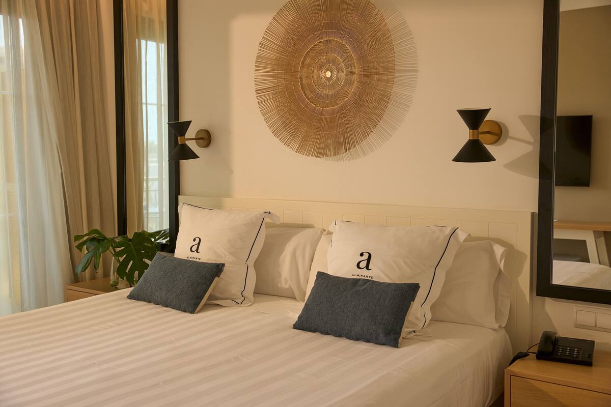 Una cama bien hecha con dos almohadas, una mesita de noche con teléfono y una lámpara de pared.