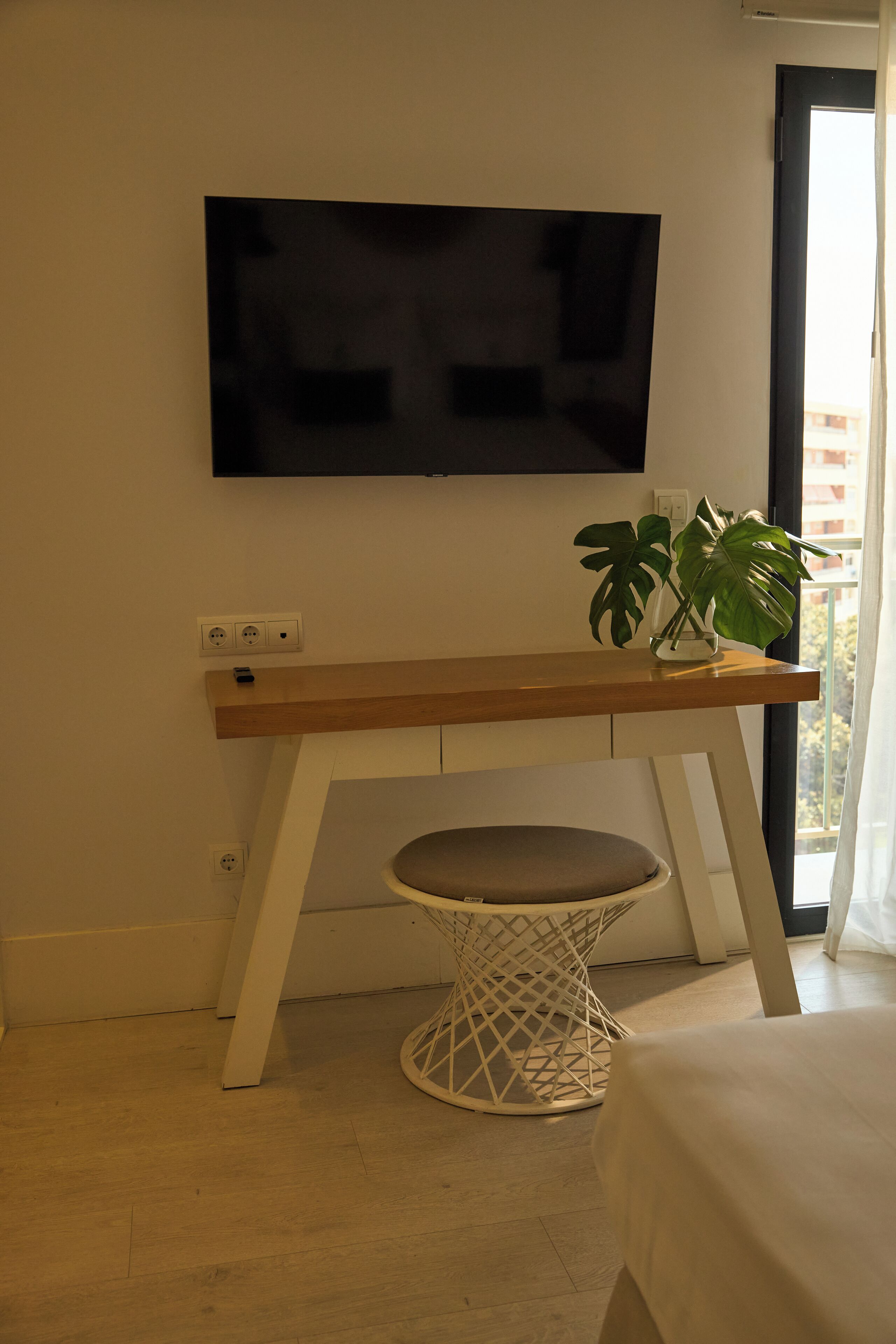 A room with a wooden desk, a white chair, a television mounted on the wall, and a potted plant.