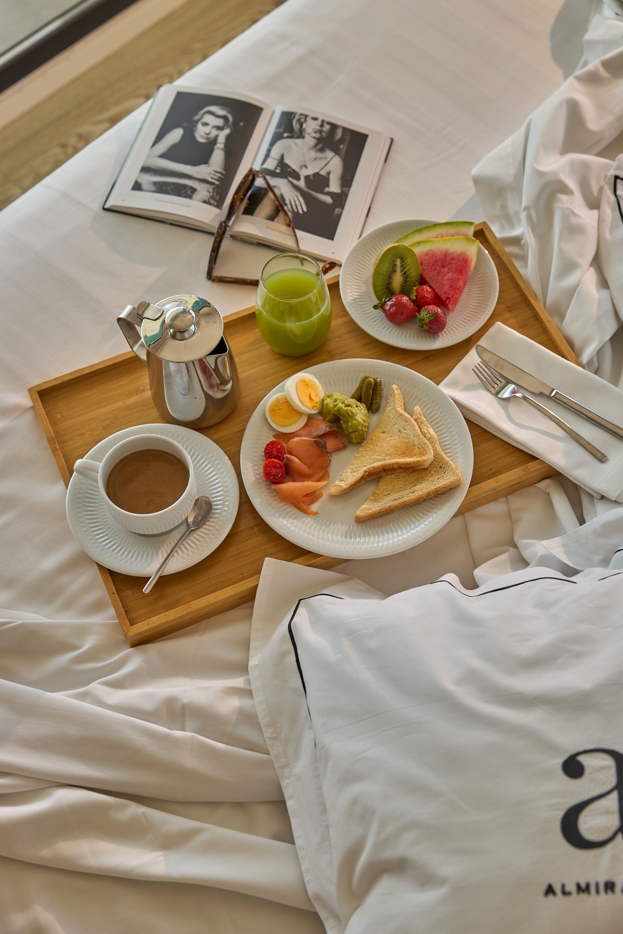 Bandeja de desayuno con café, tostadas, fruta y una bebida sobre una cama, junto a un libro y una almohada.