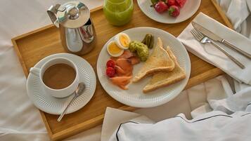 Bandeja de desayuno con café, tostadas, fruta y una bebida sobre una cama, junto a un libro y una almohada.
