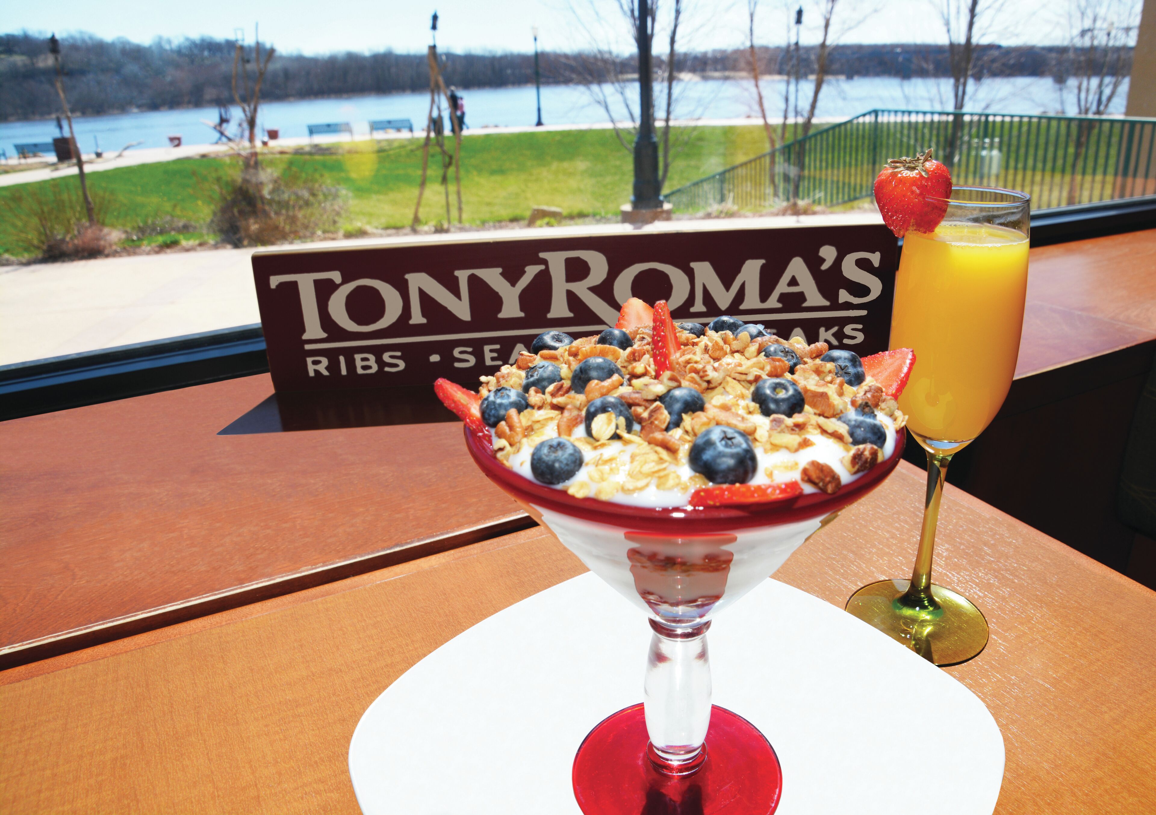 A dessert with fruit and granola on a white plate, a glass of orange juice, and a sign for Tony Roma's Ribs & Seafood in the background.