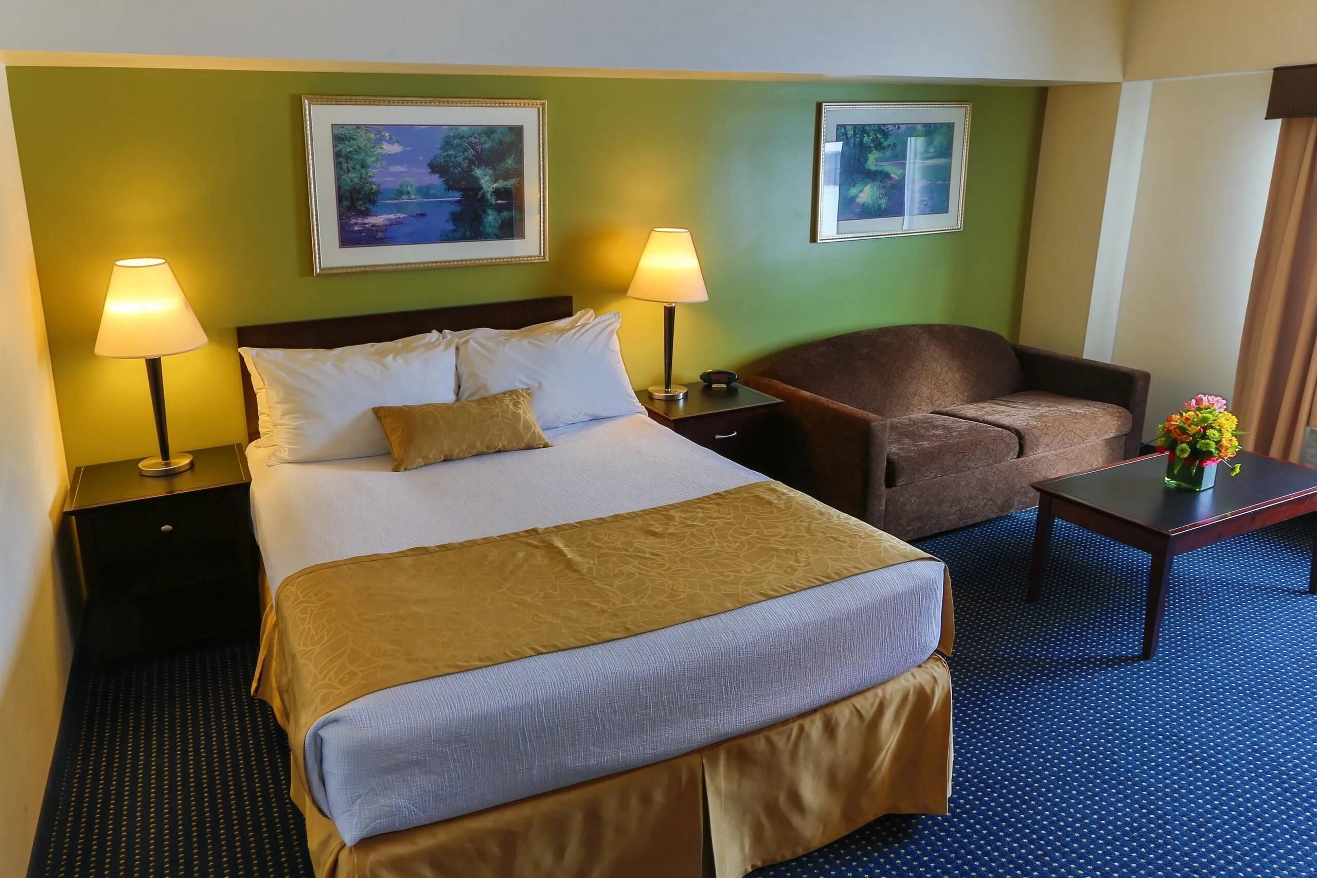A hotel room with a bed, two bedside lamps, a chair, a small table with a vase, and two framed pictures on the wall.
