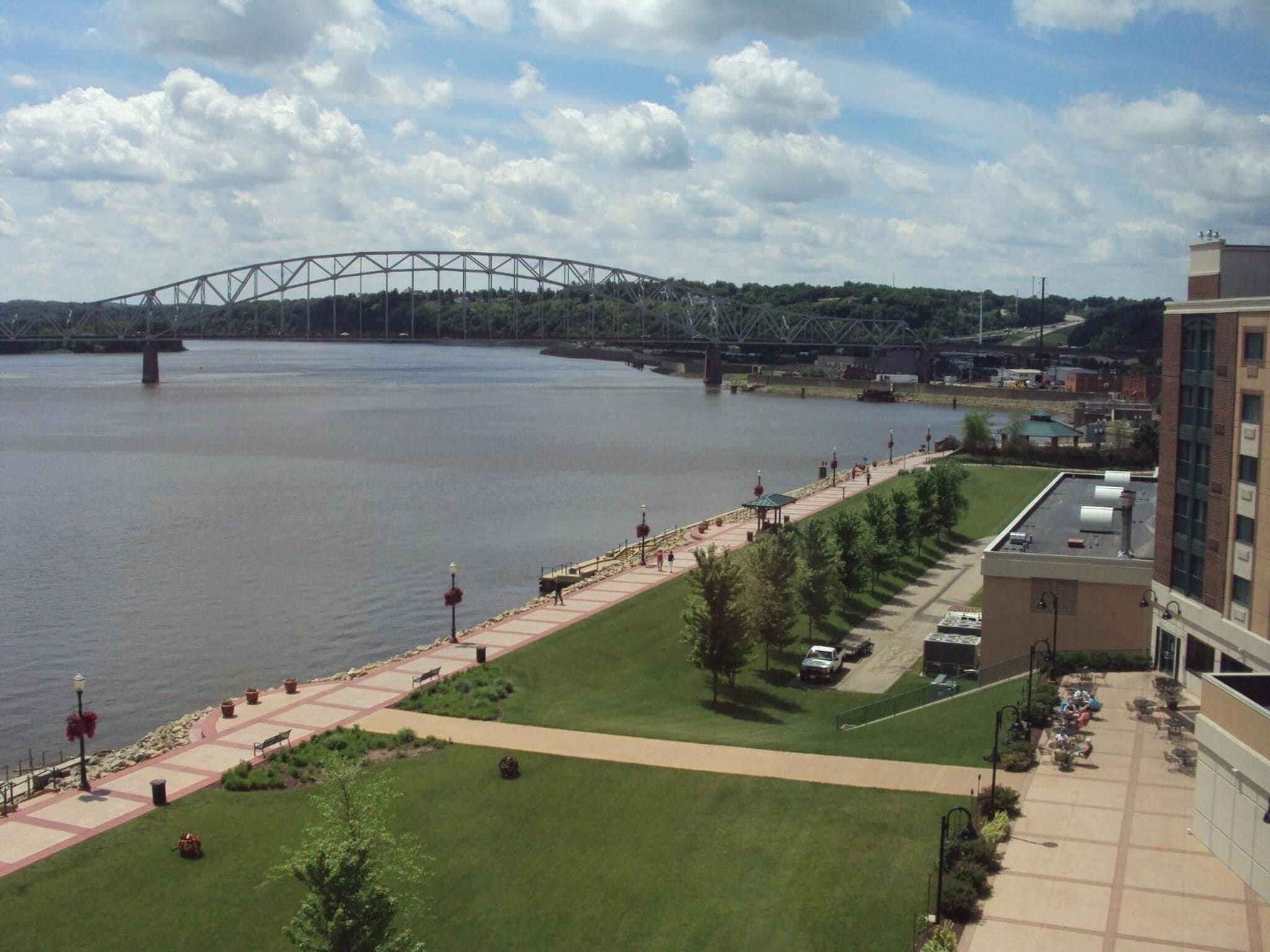 A riverside walkway with a bridge in the background, a grassy area, and a building with outdoor seating.