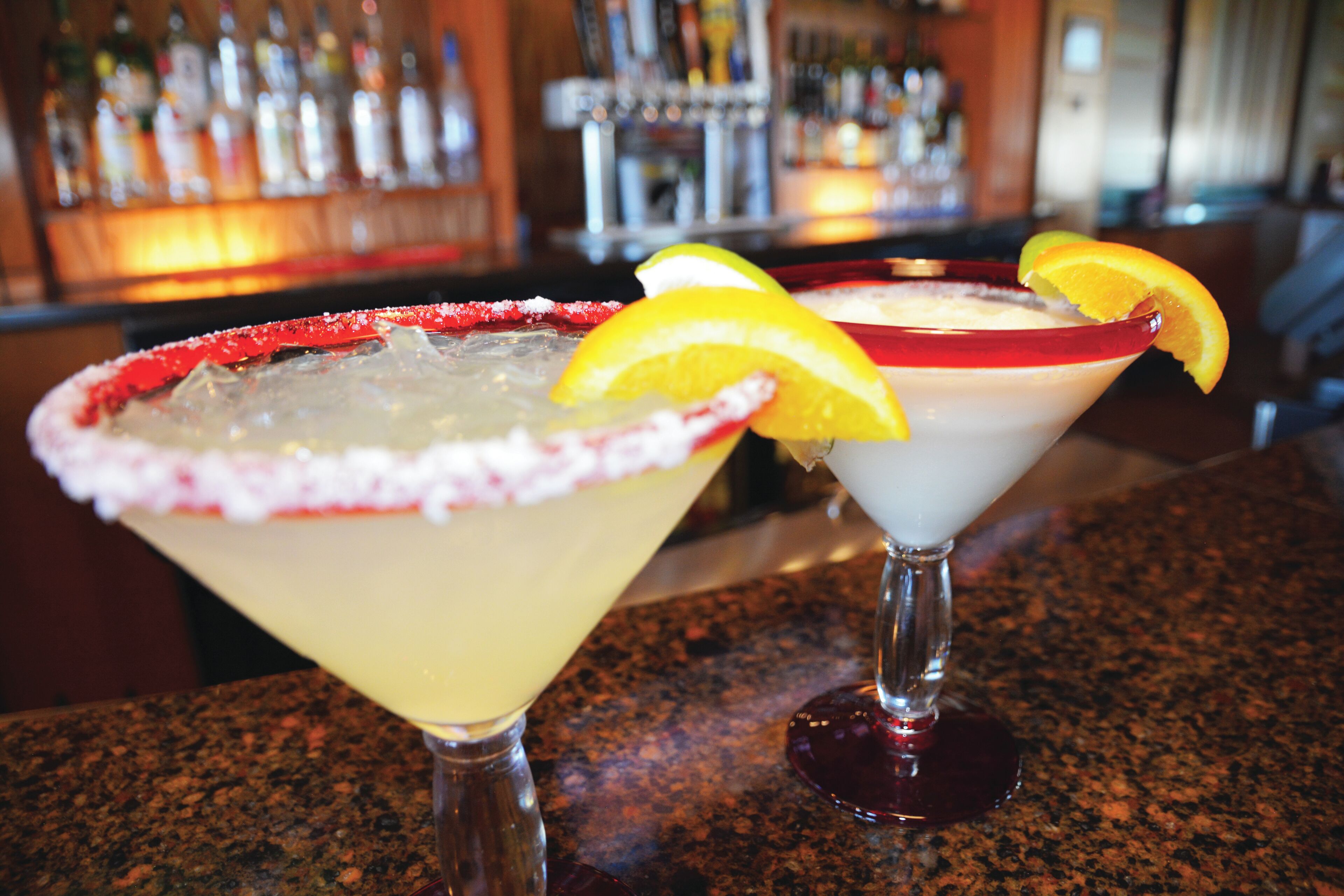 Two cocktails on a bar counter with a blurred background of bottles.