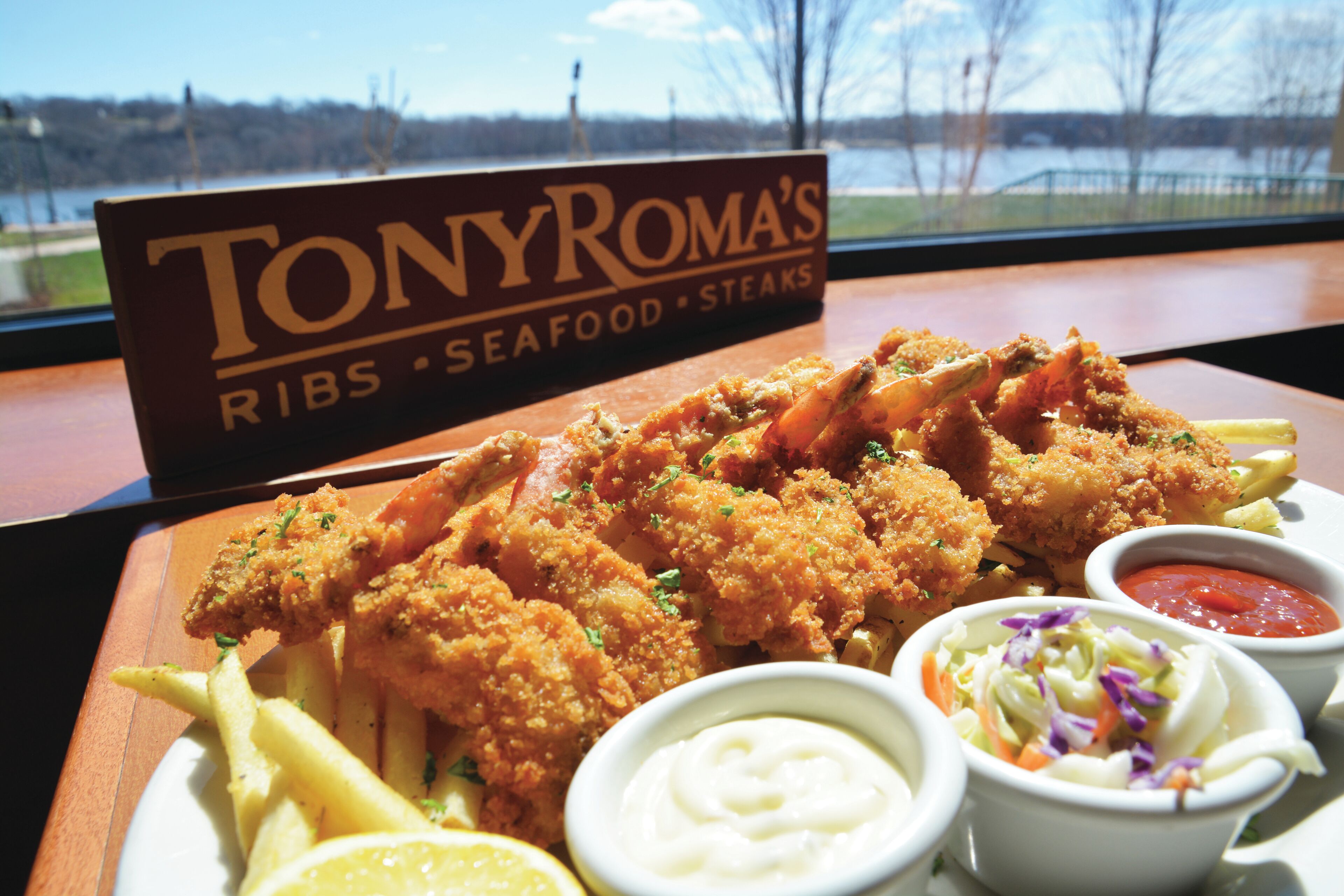 A plate of fried seafood with lemon wedges and two dipping sauces, a sign for Tony Roma's, and a view of a body of water through a window.