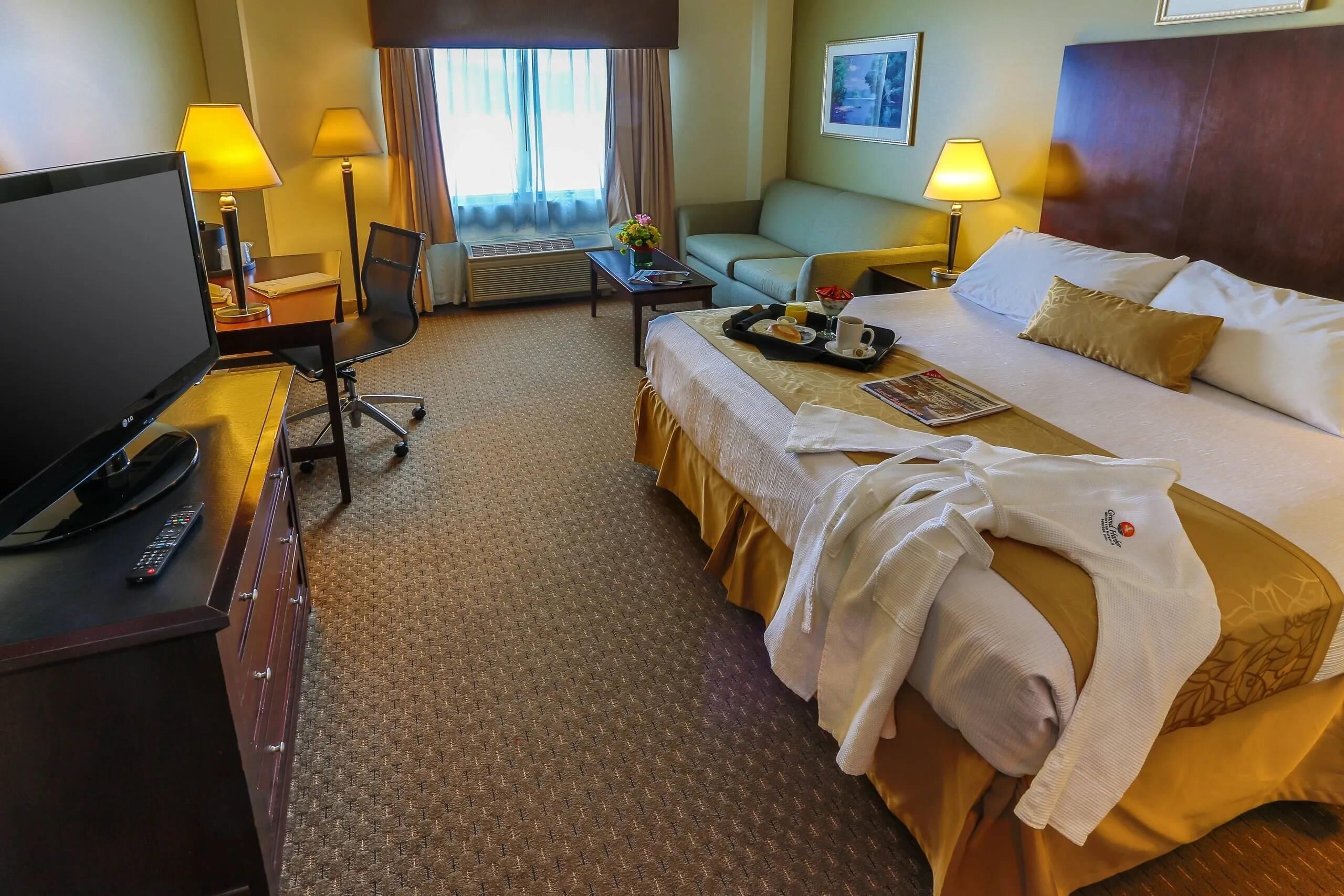 A hotel room with a bed, a desk with a TV, a chair, a sofa, and a window with curtains.
