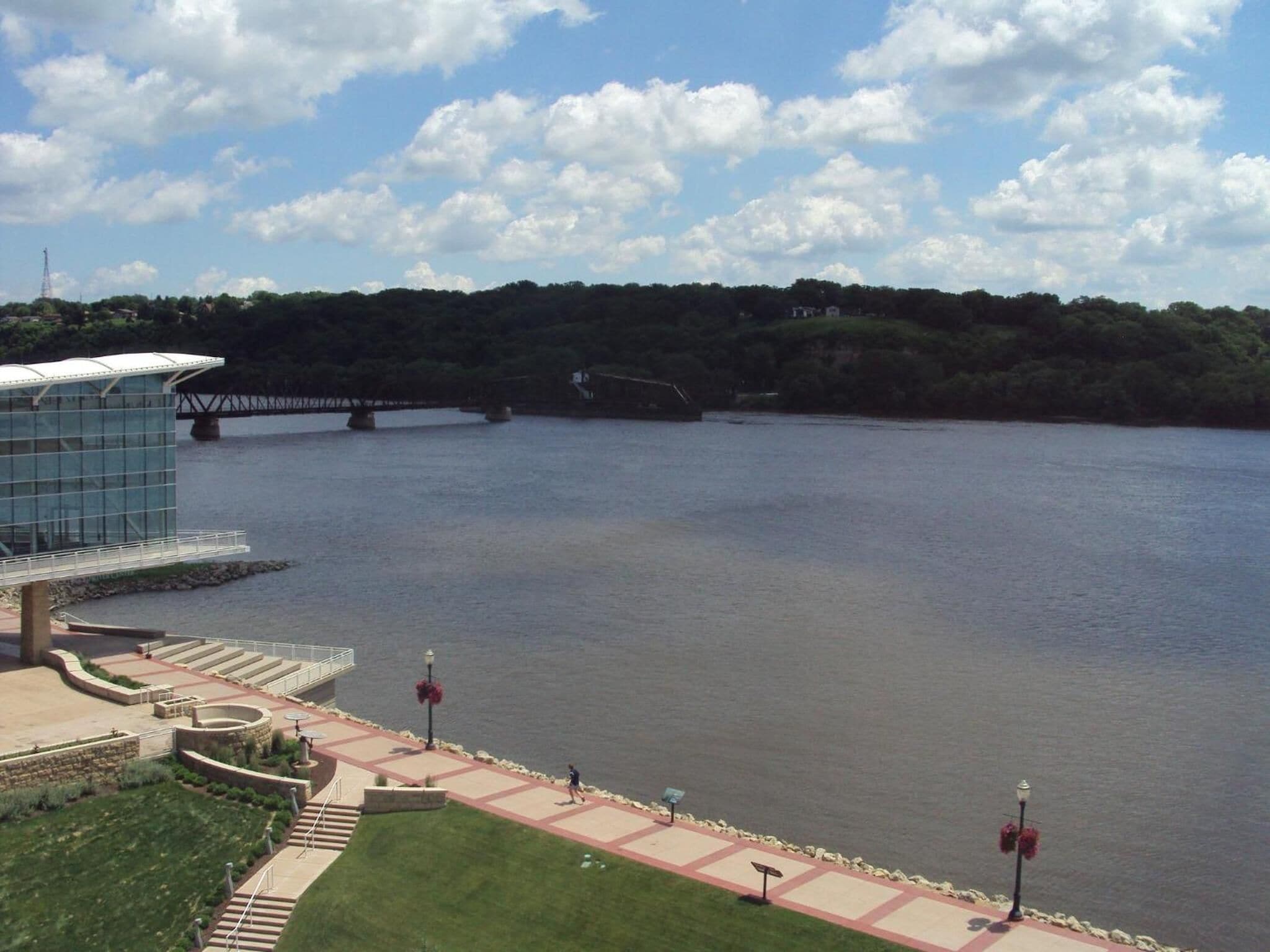 A riverside walkway with a modern building, a bridge, and a natural landscape.
