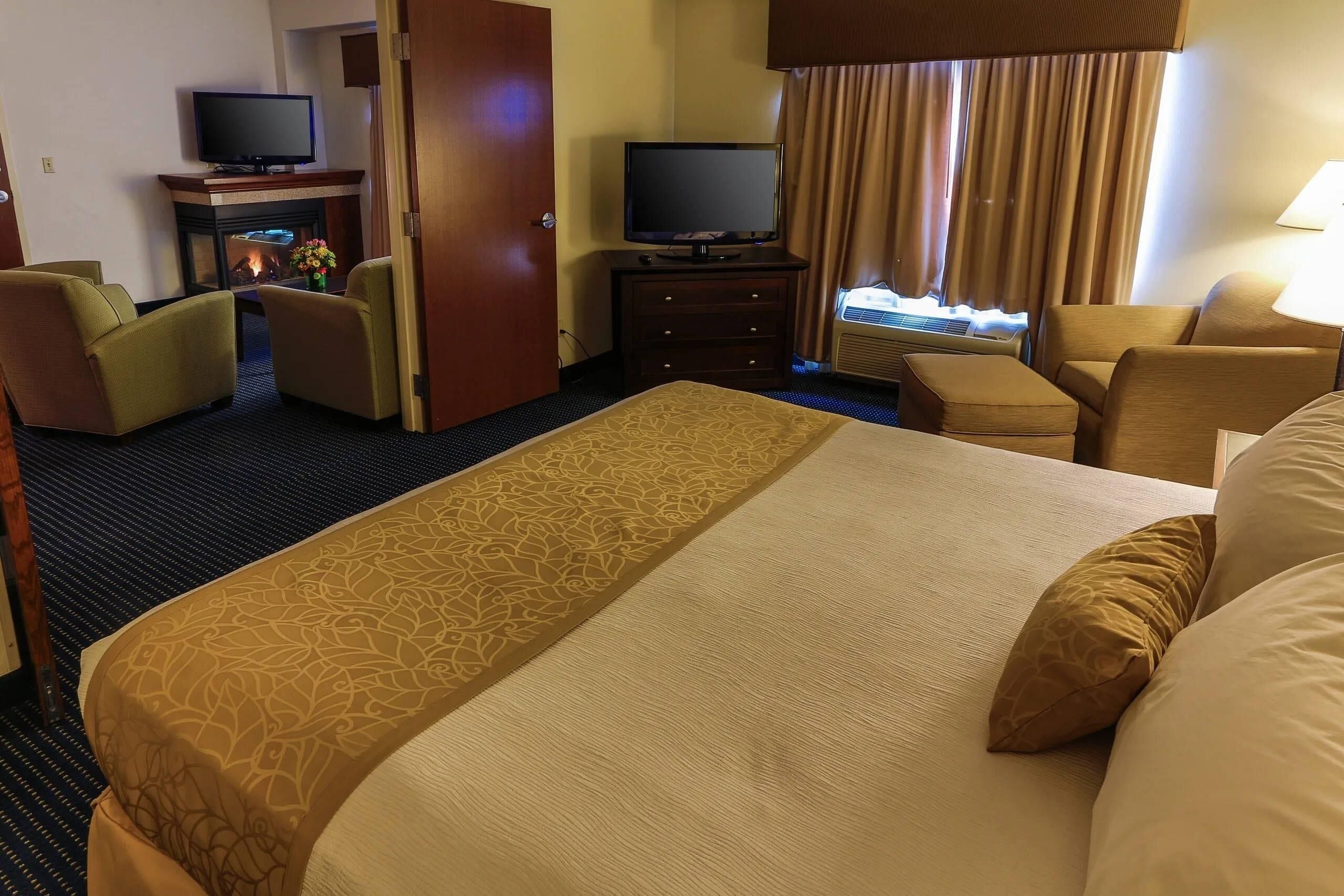 A hotel room with a large bed, two armchairs, a TV, a fireplace, and a window with curtains.