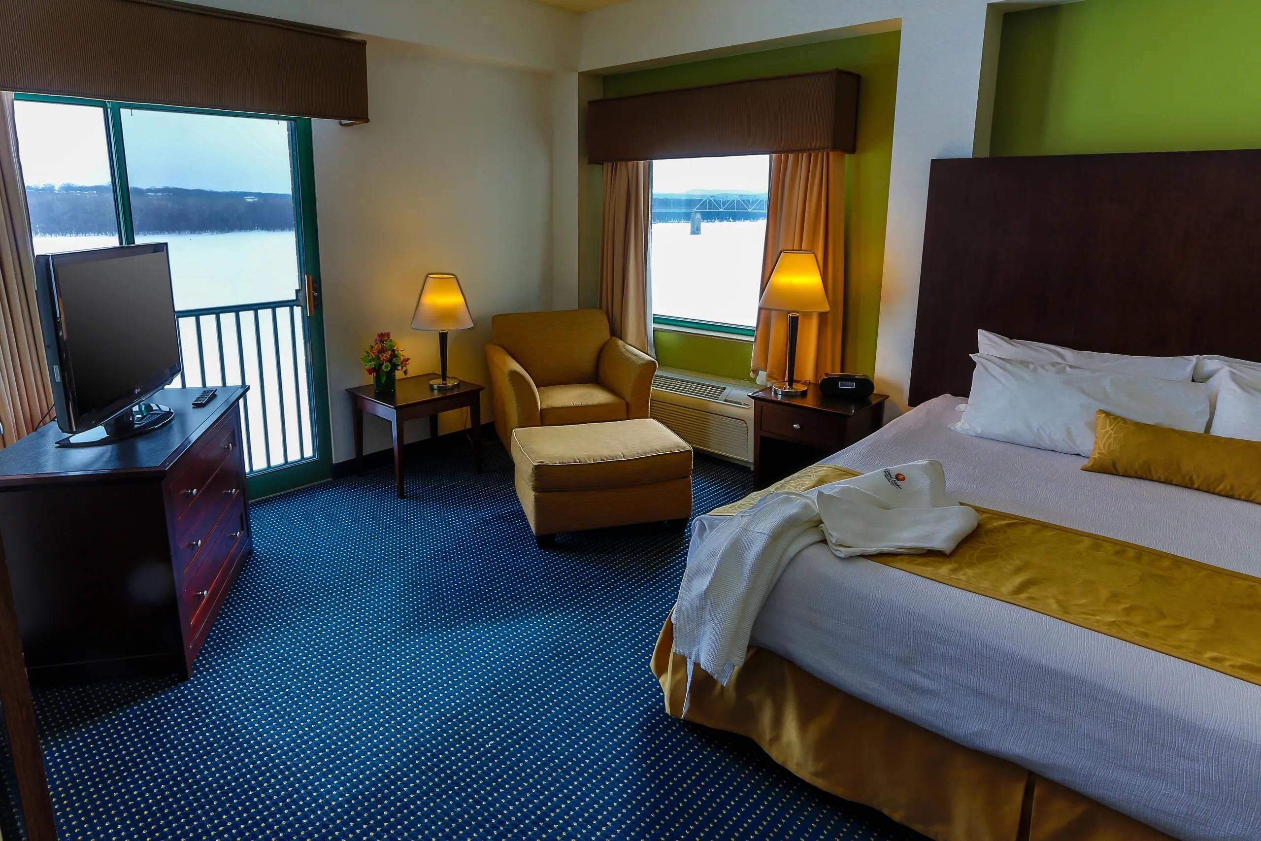 A hotel room with a bed, a chair, a desk with a TV, and a view of a body of water.