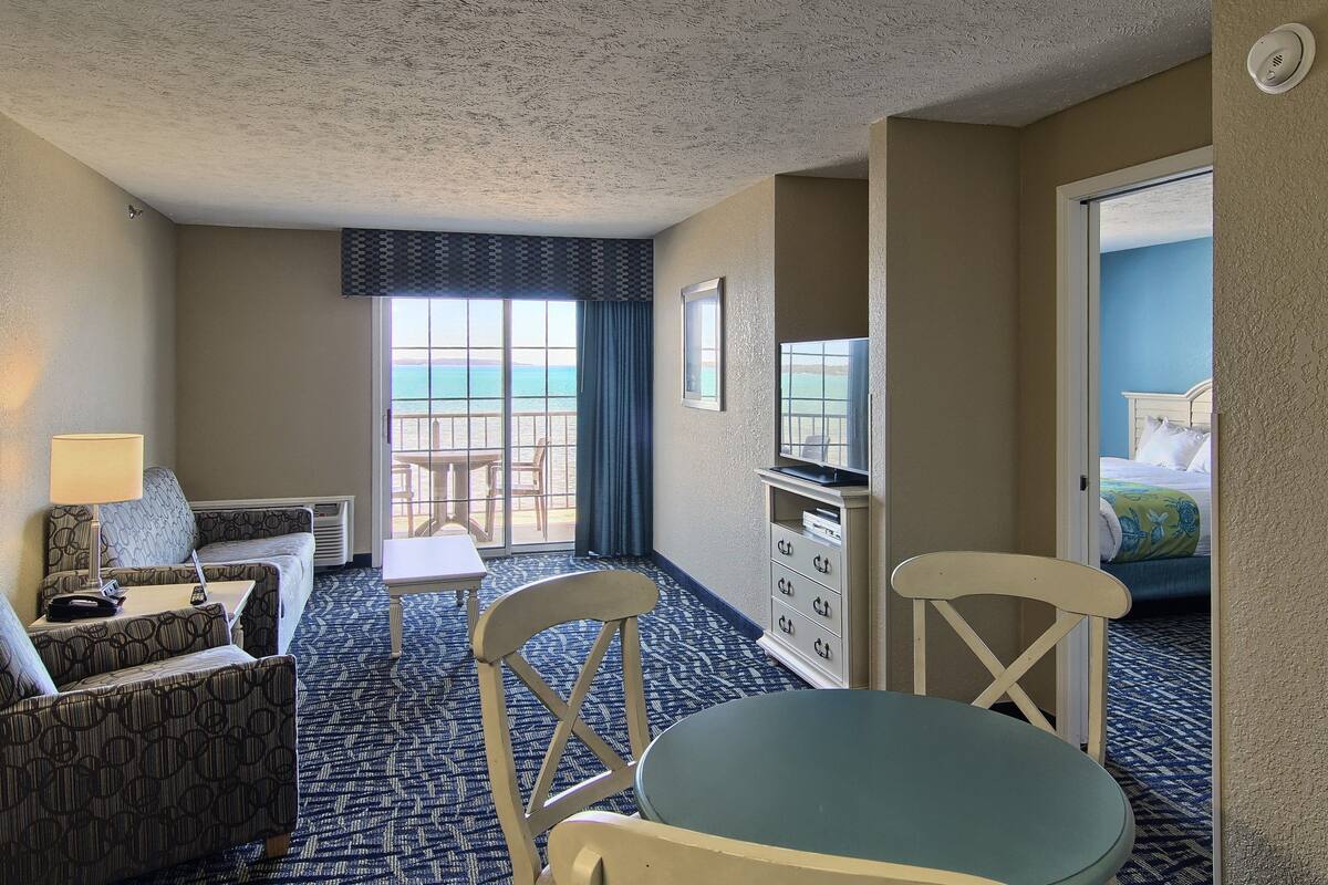 A hotel room with a balcony, a sofa, a chair, a table, and a dresser.