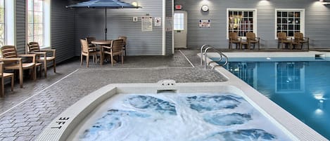 A hot tub with a tiled edge and a metal ladder, surrounded by a brick floor and wooden chairs.