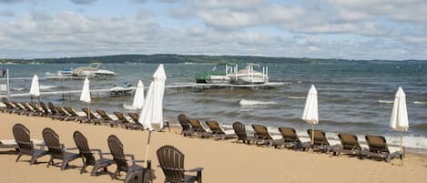 A beach with rows of sun loungers and umbrellas, a dock with boats, and a calm sea under a partly cloudy sky.