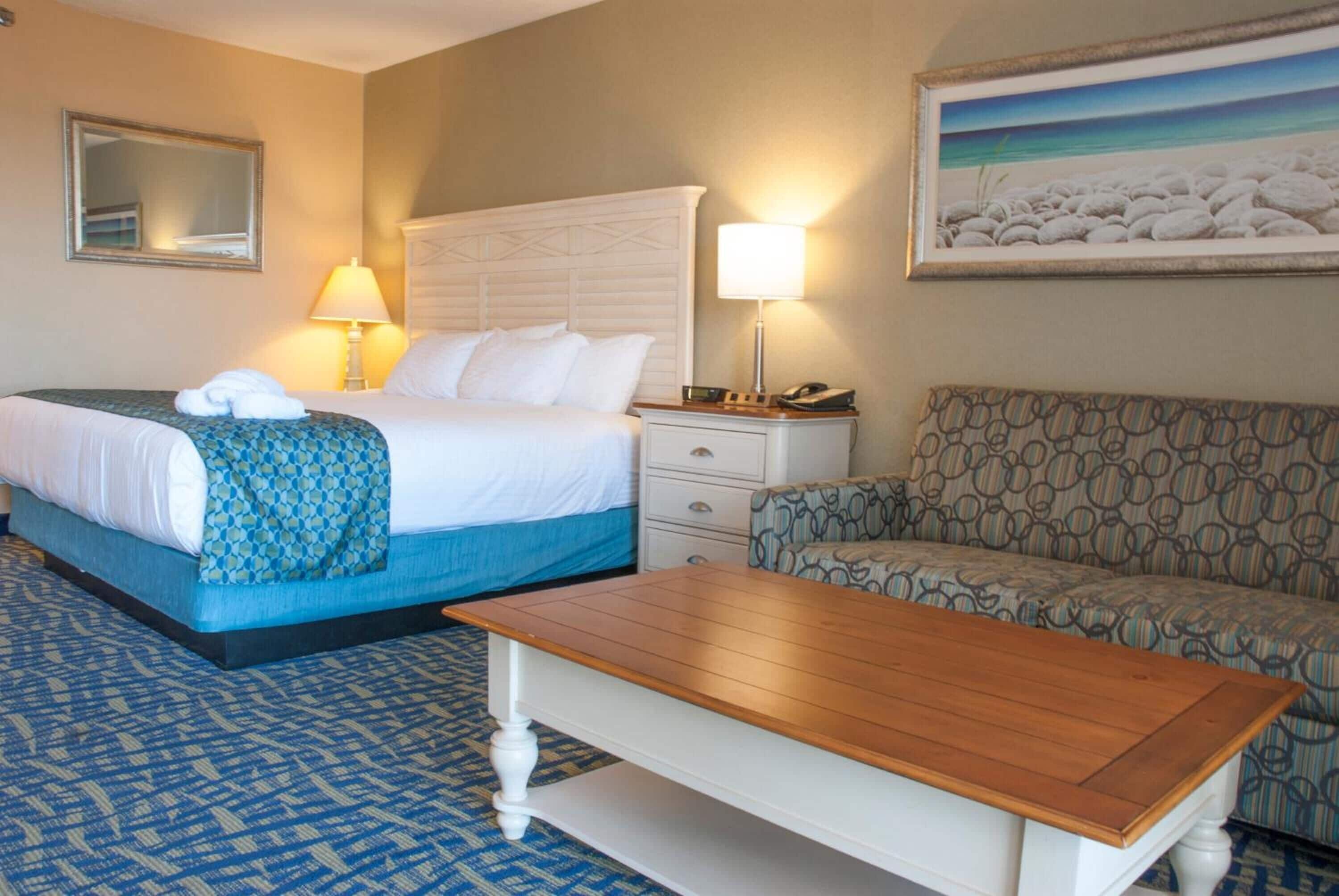 A hotel room with a bed, a bedside table with a lamp, a chair, a small table, and a painting on the wall.