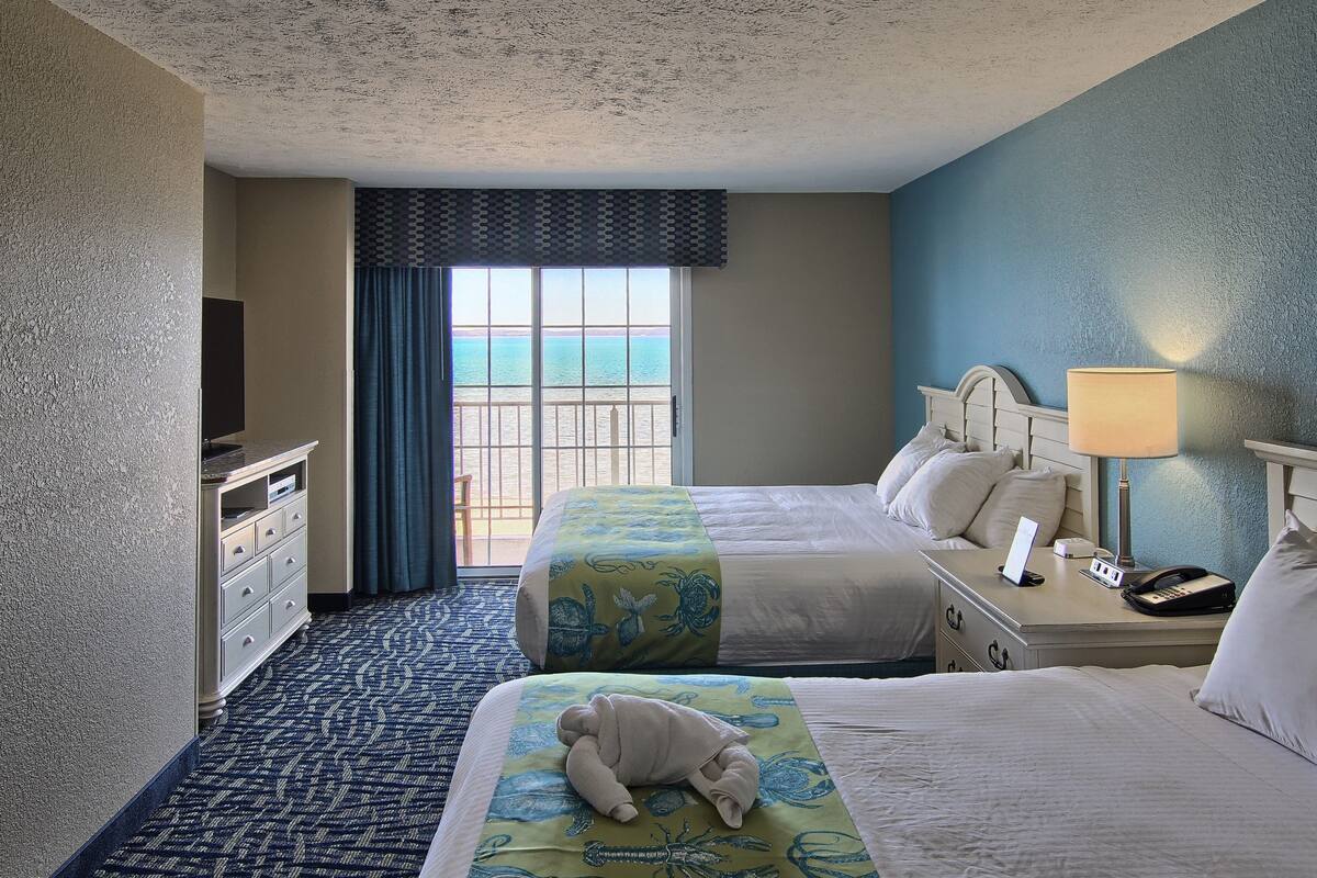 A hotel room with two beds, a dresser, a desk with a lamp, and a view of the ocean through a large window.