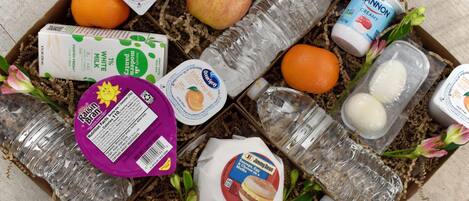 Variedad de alimentos y bebidas dispuestas en una caja, entre ellas un cartón de leche, una botella de jugo, un paquete de muffins y varias manzanas.