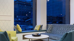 A modern living room with a grey sofa, teal armchairs, and a coffee table. Large windows offer a view of a cityscape at dusk. - Hotel Capitol (Bucharest)