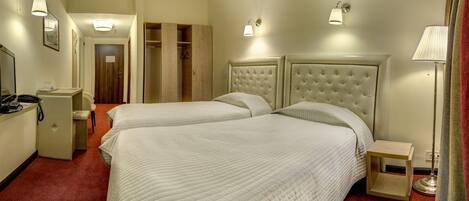A hotel room with two beds, a desk, a chair, a wardrobe, and a bedside table with a lamp.