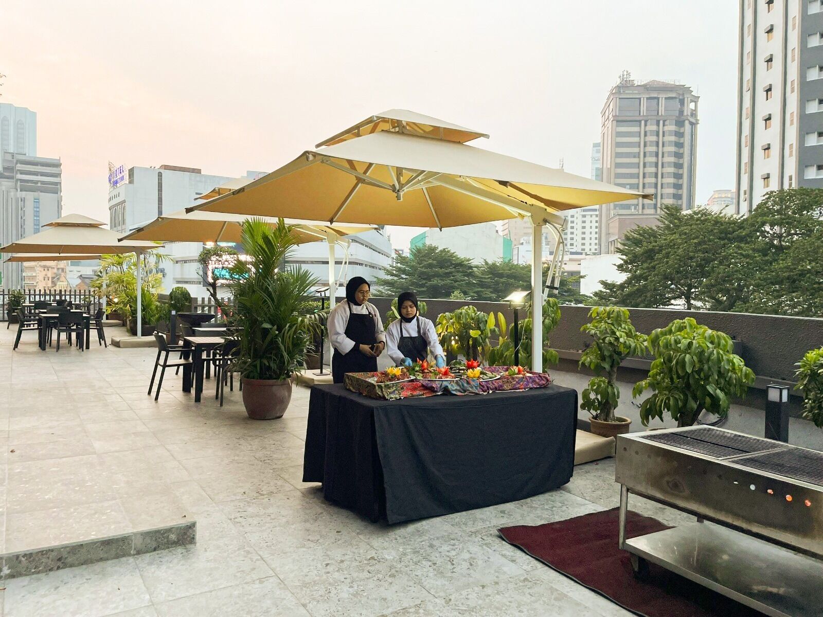 An outdoor dining area with tables and chairs, a grill station, and umbrellas providing shade.