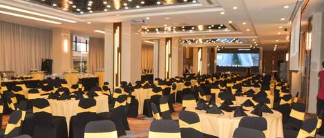 A conference room with rows of tables and chairs, a stage with a screen, and a speaker at the front.