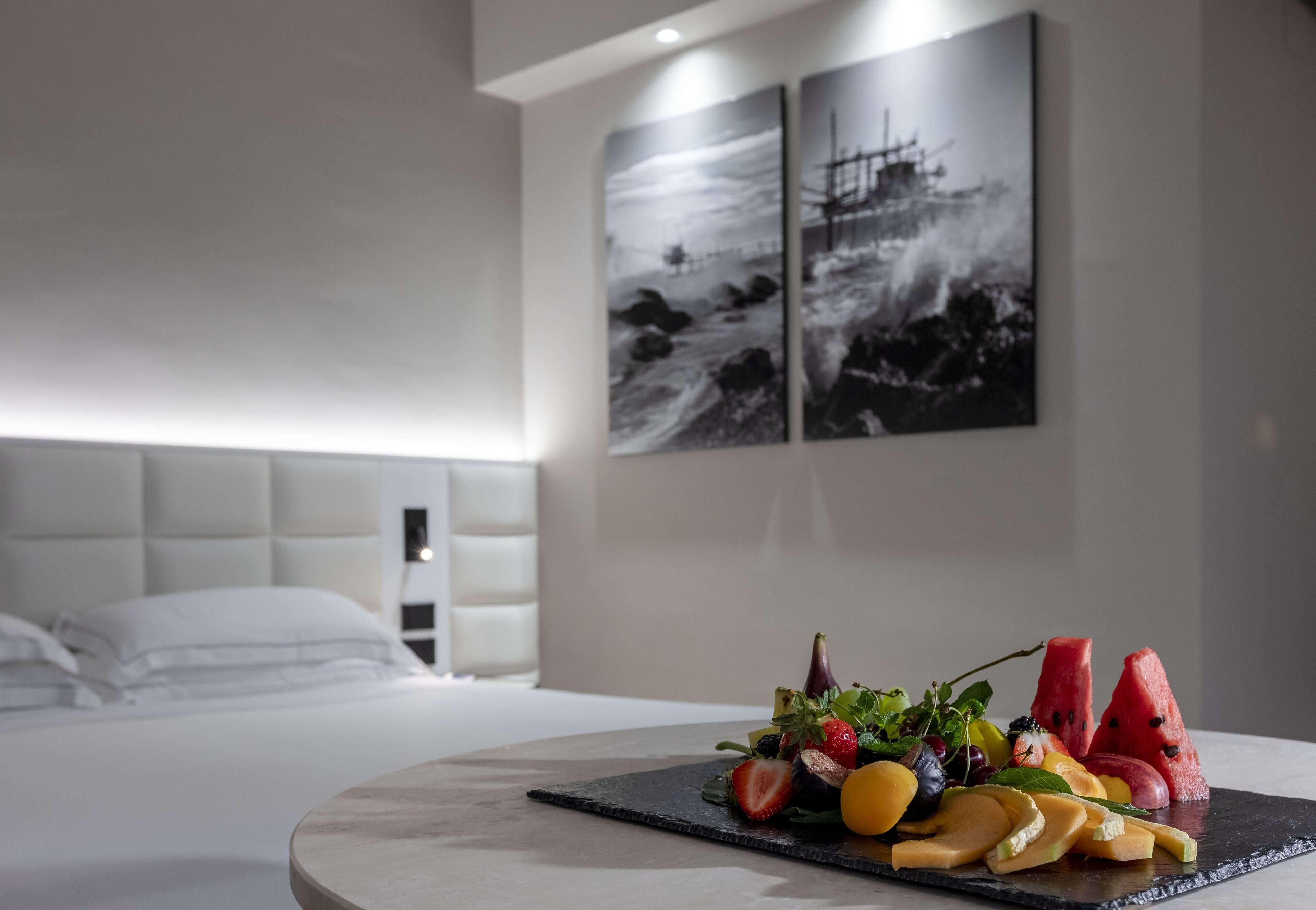 A hotel room with a bed, a fruit platter on a marble table, and two black-and-white maritime paintings on the wall.