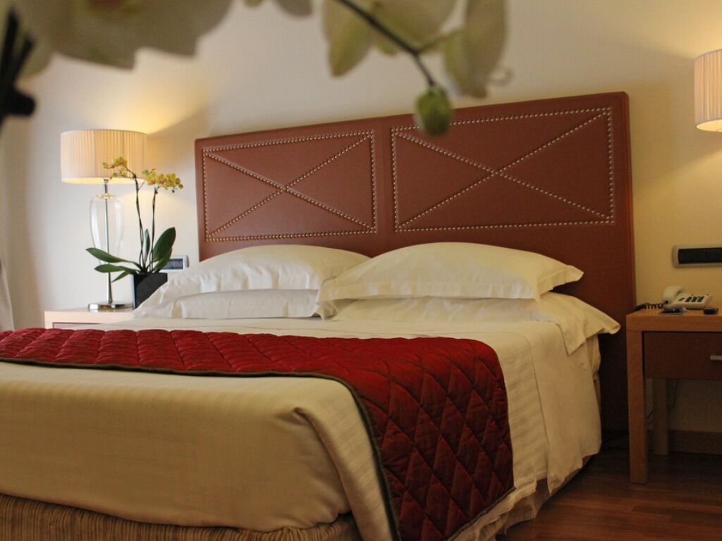 Un lit bien fait, recouvert d’une couverture rouge, avec une tête de lit en bois ornée d’un motif géométrique, une lampe de chevet et une petite table sur laquelle se trouve un téléphone.