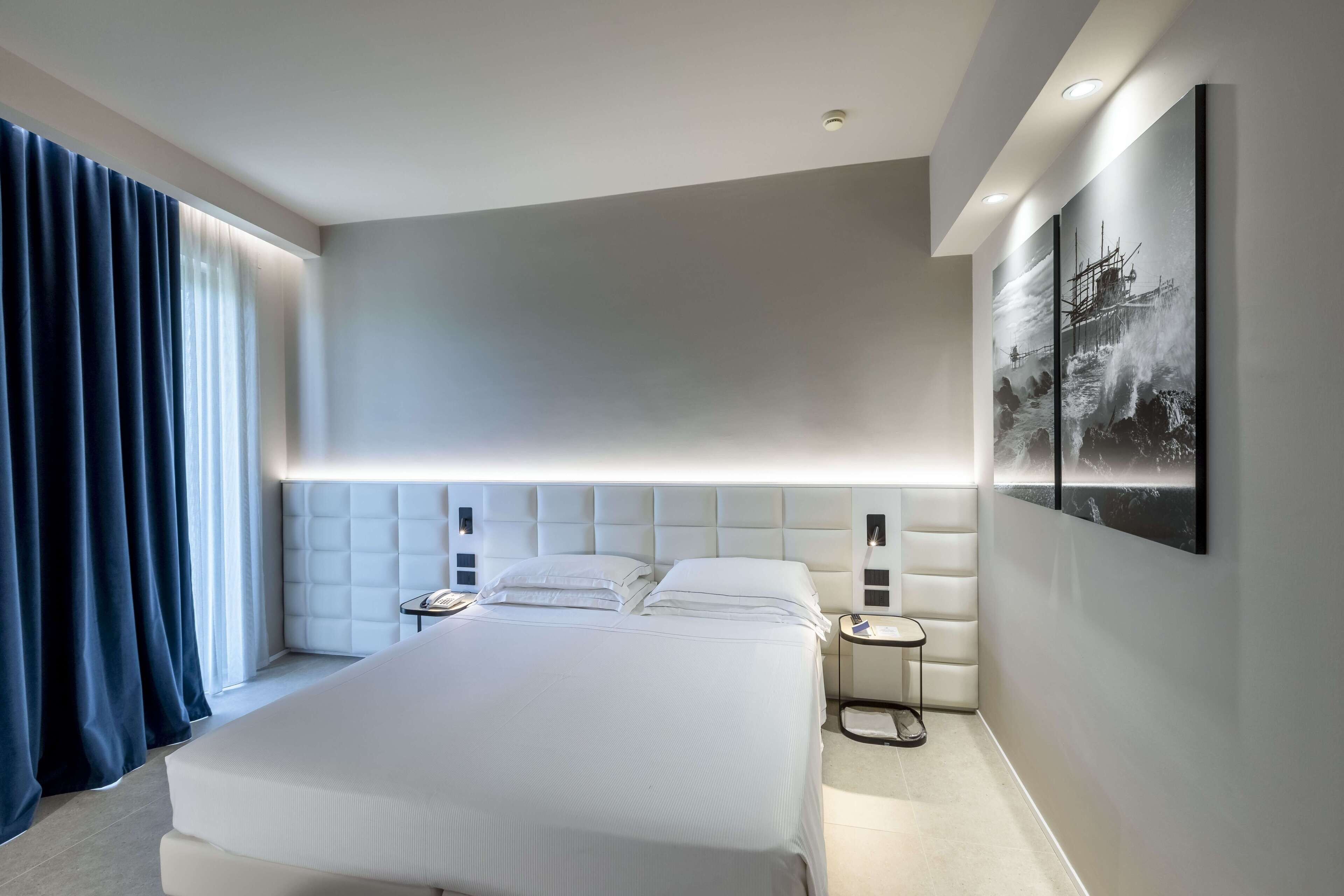Camera d'albergo moderna con un letto grande, una testiera illuminata e un comodino con un telefono, oltre a un'opera d'arte appesa al muro.