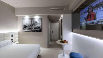 A hotel room with a bed, a small table with fruit, a TV screen displaying news, and a wall with two black-and-white paintings.