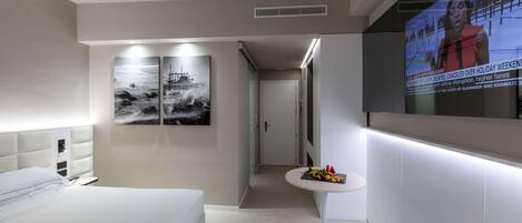 A hotel room with a bed, a small table with fruit, a TV screen displaying news, and a wall with two black-and-white paintings.