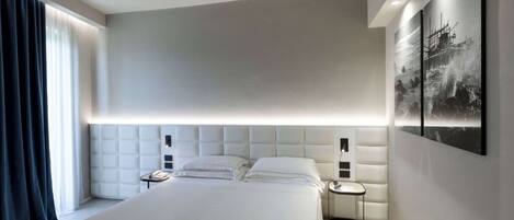 Quarto de hotel moderno com uma cama grande, cabeceira com iluminação integrada, mesa de cabeceira com telefone e uma obra de arte pendurada na parede.