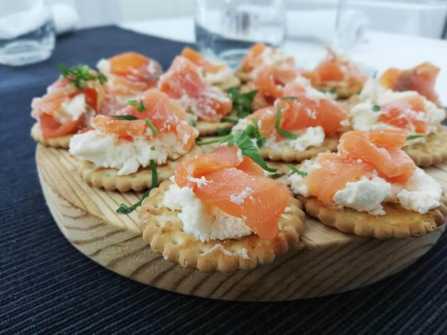 Una tabla de madera con varios aperitivos, entre ellos galletas cubiertas de queso crema y salmón ahumado, adornada con hierbas.