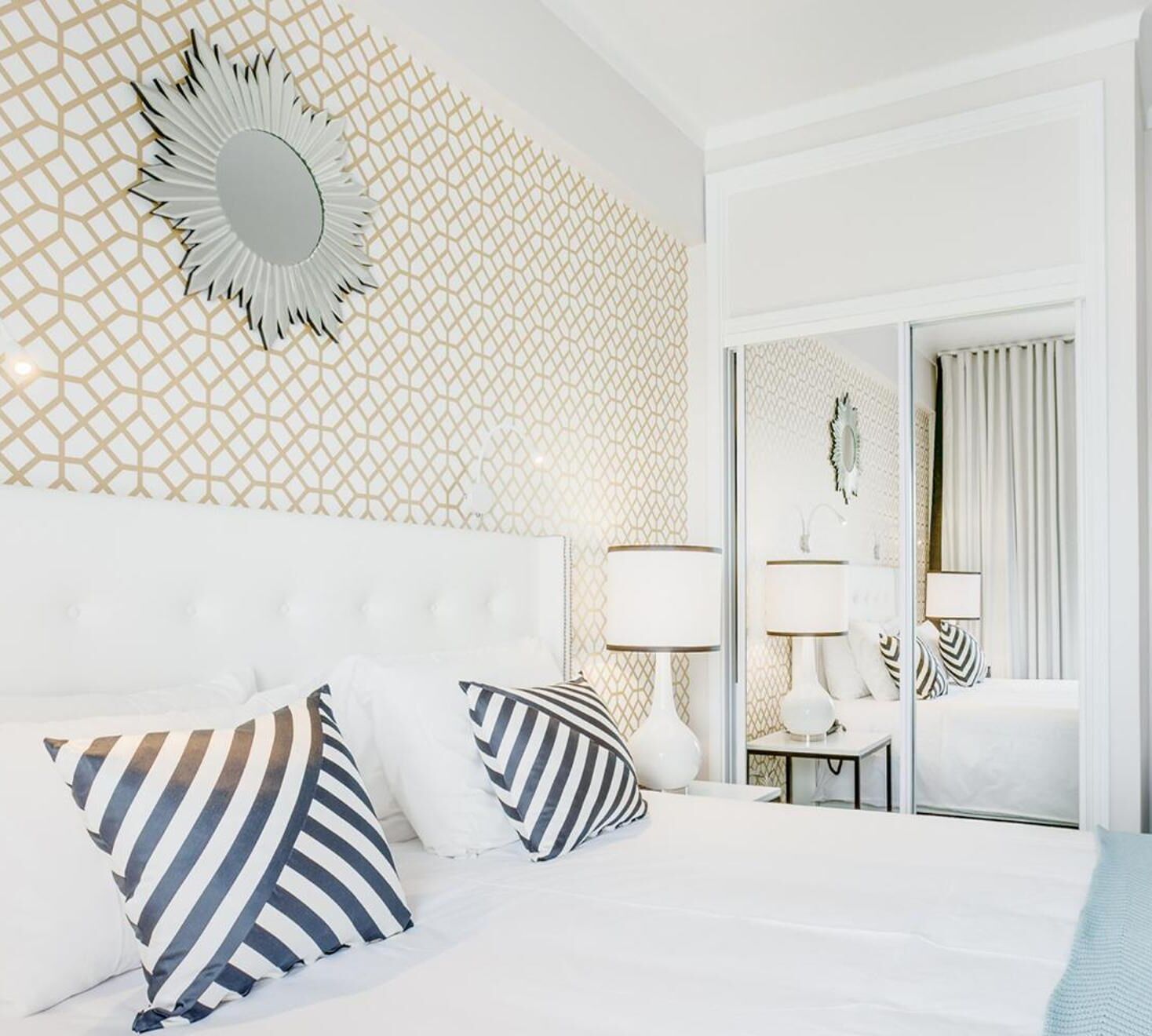 Ein Hotelzimmer mit einem weißen Bett, gestreifteren Kissen, einem Spiegel mit Sonnenstrahlendesign und einer Wand mit geometrischem Muster.