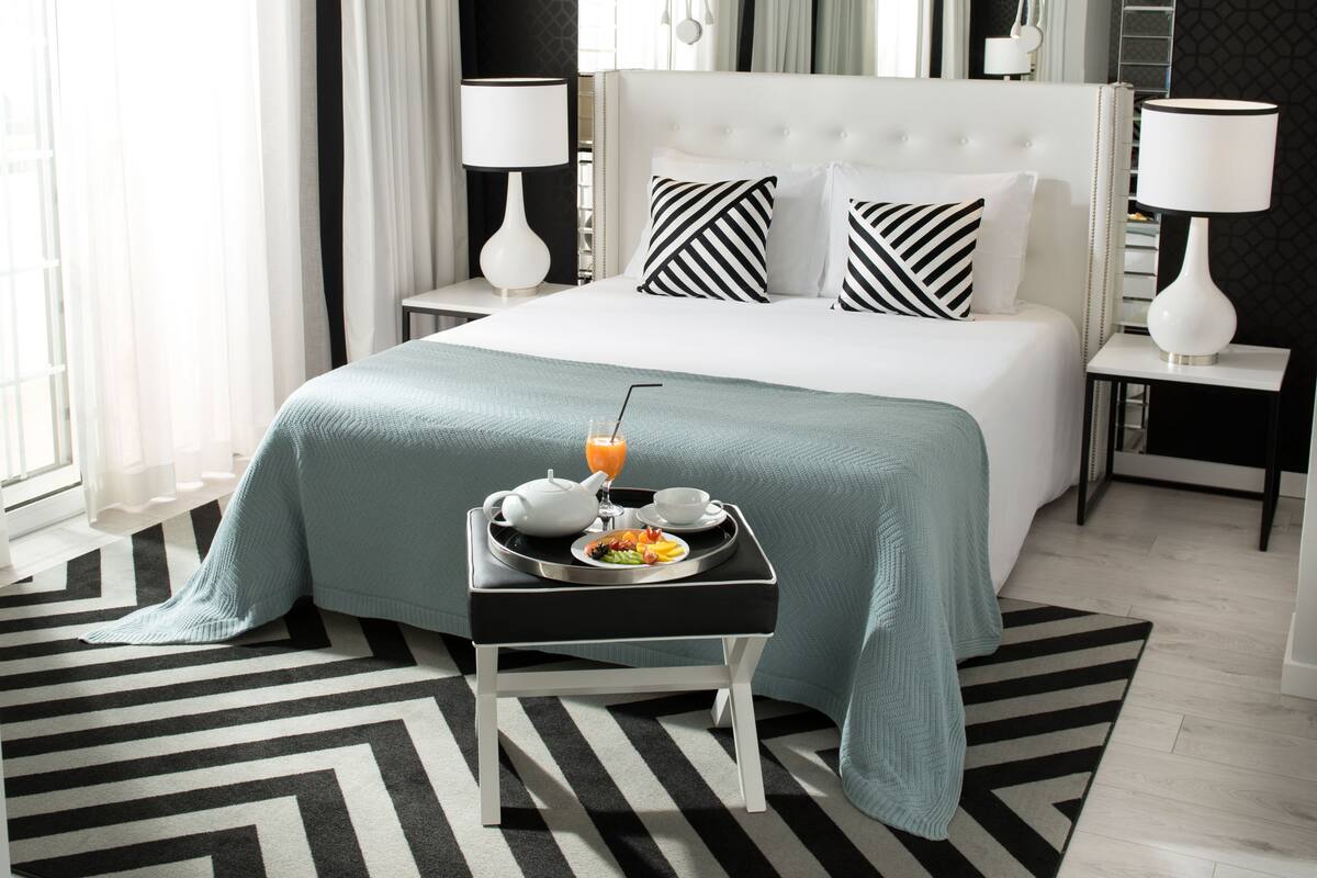 Una habitación de hotel moderna con una cama grande, una mesita de noche con una bandeja de desayuno y vistas a la ciudad desde la ventana.