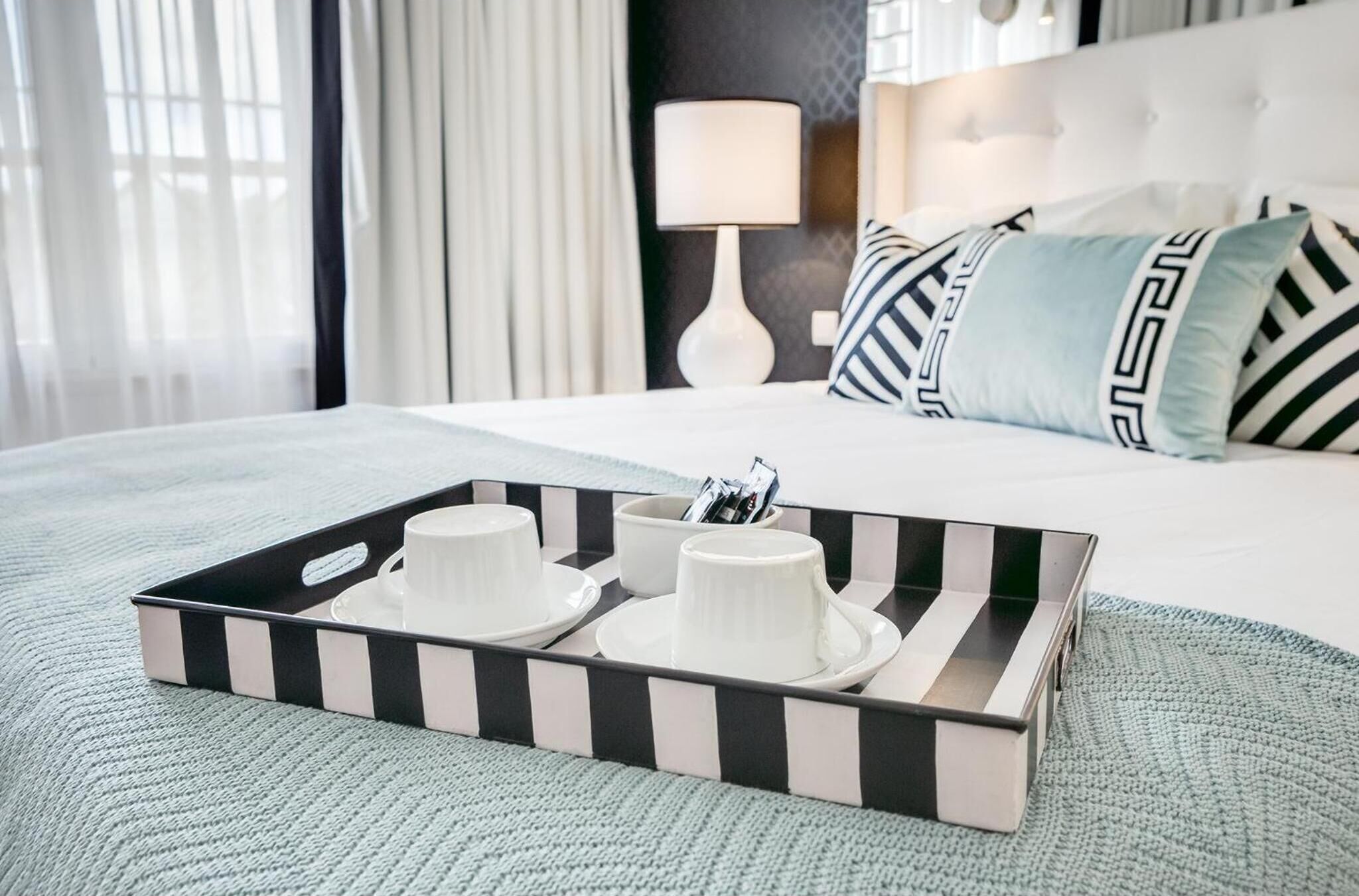 A neatly made bed with a tray of white cups and saucers, a lamp, and patterned pillows.