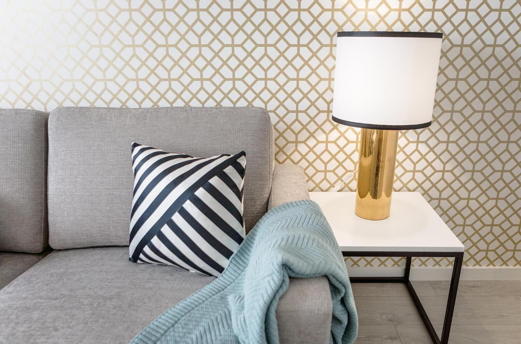 A modern living room with a grey sofa, a striped pillow, a white table with a gold lamp, and patterned wallpaper.