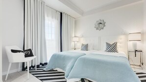 A modern hotel room with two beds, a large window with white curtains, a black and white striped rug, and a white chair with a pillow. - Hotel Cristal Marinha (Marinha Grande)