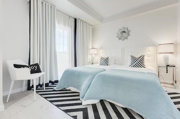 Una habitación de hotel moderna con dos camas, un ventanal con cortinas blancas, un tapete a rayas blanco y negro y una silla blanca con un cojín.