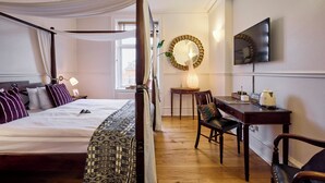 Un lit à baldaquin avec du linge de lit blanc, un bureau en bois avec une chaise, un miroir rond et un téléviseur à écran plat fixé au mur.