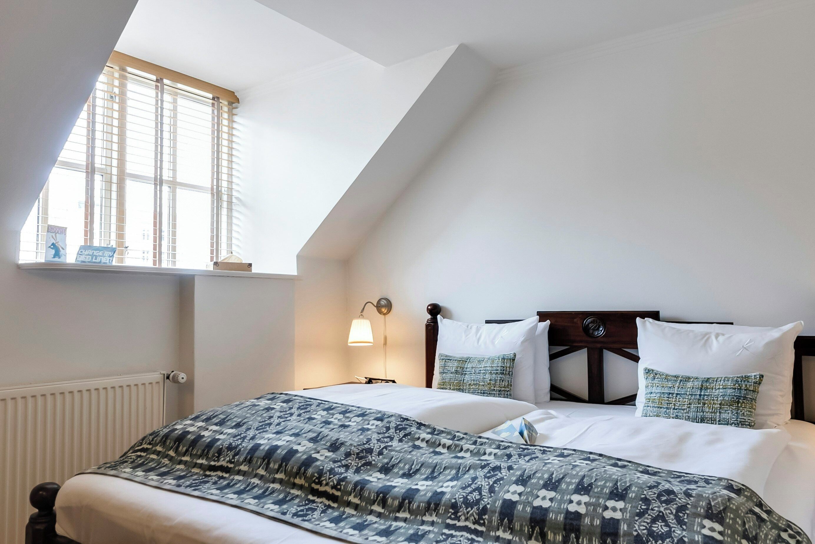 Ein Schlafzimmer mit schrägem Dach, einem Fenster mit Jalousien, einer Nachttischlampe, einem Bett mit gemusterten Betttextilien und einem Zierdekor an der Kopfteil.