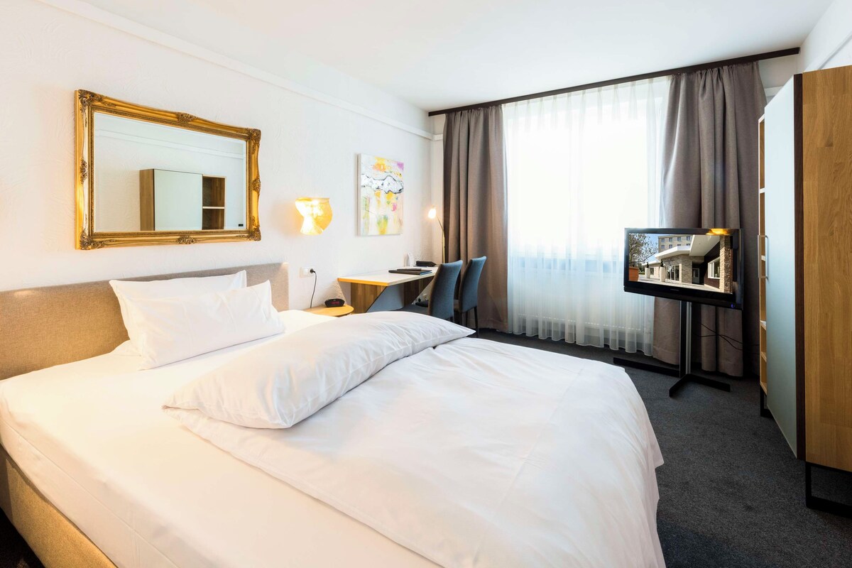 A hotel room with a large bed, a desk, a chair, a TV, and a window with curtains.