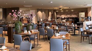 Un restaurante moderno con varias mesas y sillas, una planta decorativa en el centro y una zona de cocina al fondo.