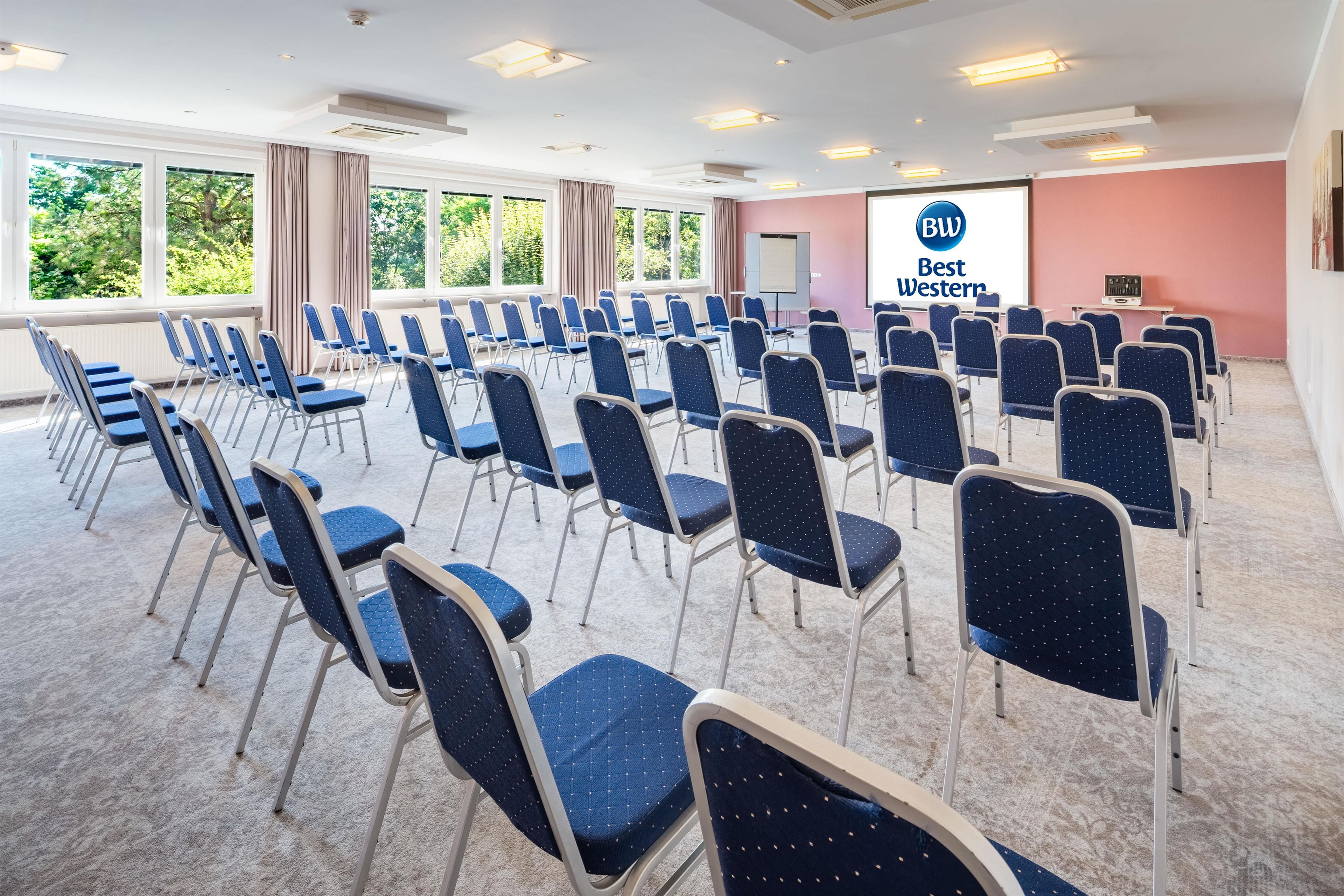 Un salón de conferencias con filas de sillas, una pantalla de proyector y un ventanal con vista a árboles.